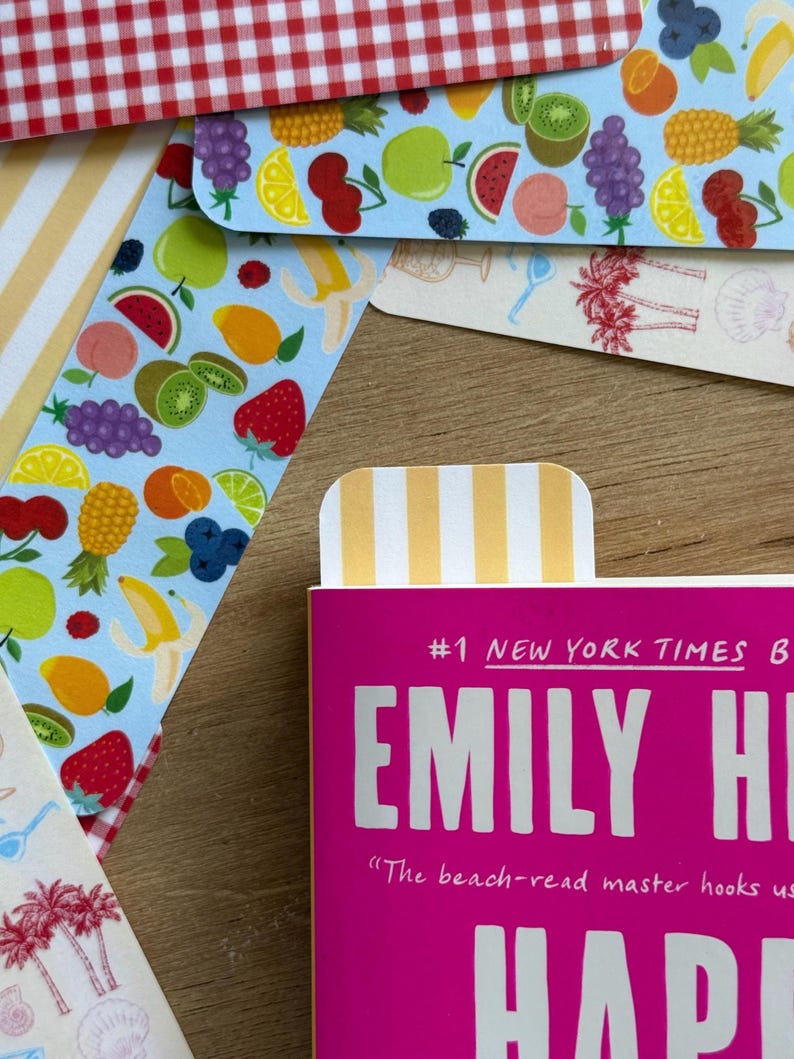 May include: A collection of colorful bookmarks with fruit and striped patterns. One bookmark is inserted into a pink book with the text "#1 NEW YORK TIMES B EMILY HI". Other bookmarks feature illustrations of fruits like strawberries, bananas, and grapes.