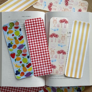 May include: A collection of colorful bookmarks. One features a red and white gingham pattern, another has yellow and white stripes, and others display fruit and tropical designs. The bookmarks are placed in an open book.