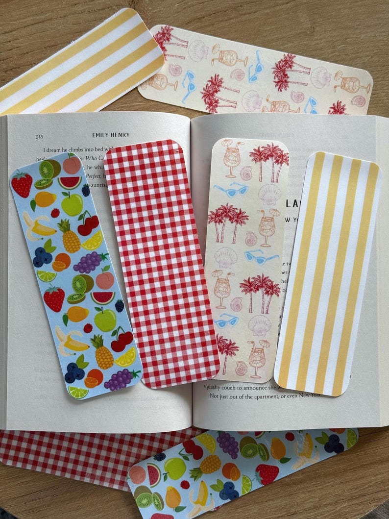 May include: A collection of colorful bookmarks. One features a red and white gingham pattern, another has yellow and white stripes, and others display fruit and tropical designs. The bookmarks are placed in an open book.