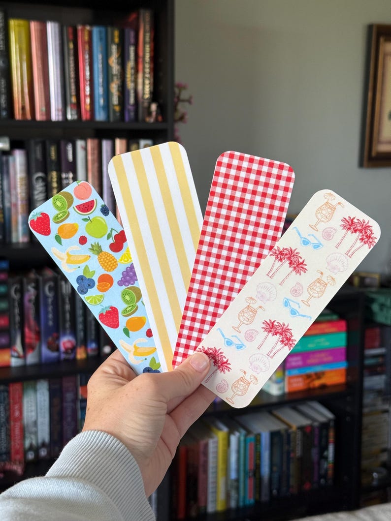 May include: Four colorful bookmarks are held in a hand. One features a fruit pattern, another has yellow stripes, a third has a red gingham pattern, and the last has a tropical design with palm trees and cocktails. Bookshelves are in the background.