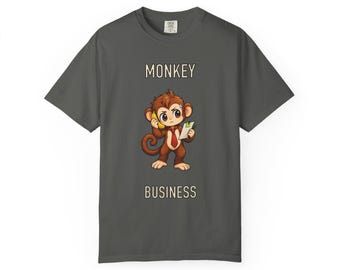 Op de afbeelding: Een antracietgrijs T-shirt met korte mouwen en een cartoon-aap met een rode das, die een banaan als telefoon tegen het oor houdt en een klembord in de andere hand heeft. De tekst MONKEY BUSINESS staat in crème boven en onder de afbeelding.