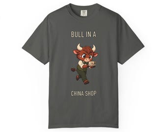 Op de afbeelding: Een antracietgrijs t-shirt met een cartoonillustratie van een bruine stier die een vest draagt en een theekopje vasthoudt. De tekst boven de stier luidt BULL IN A en de tekst eronder luidt CHINA SHOP. De stier heeft grote ogen en hoorns.
