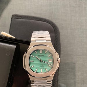 May include: A stainless steel Seiko watch with a light green face and a date display at the 3 o'clock position. The watch has a silver band and the word "SEIKO" is printed at the top of the watch face.