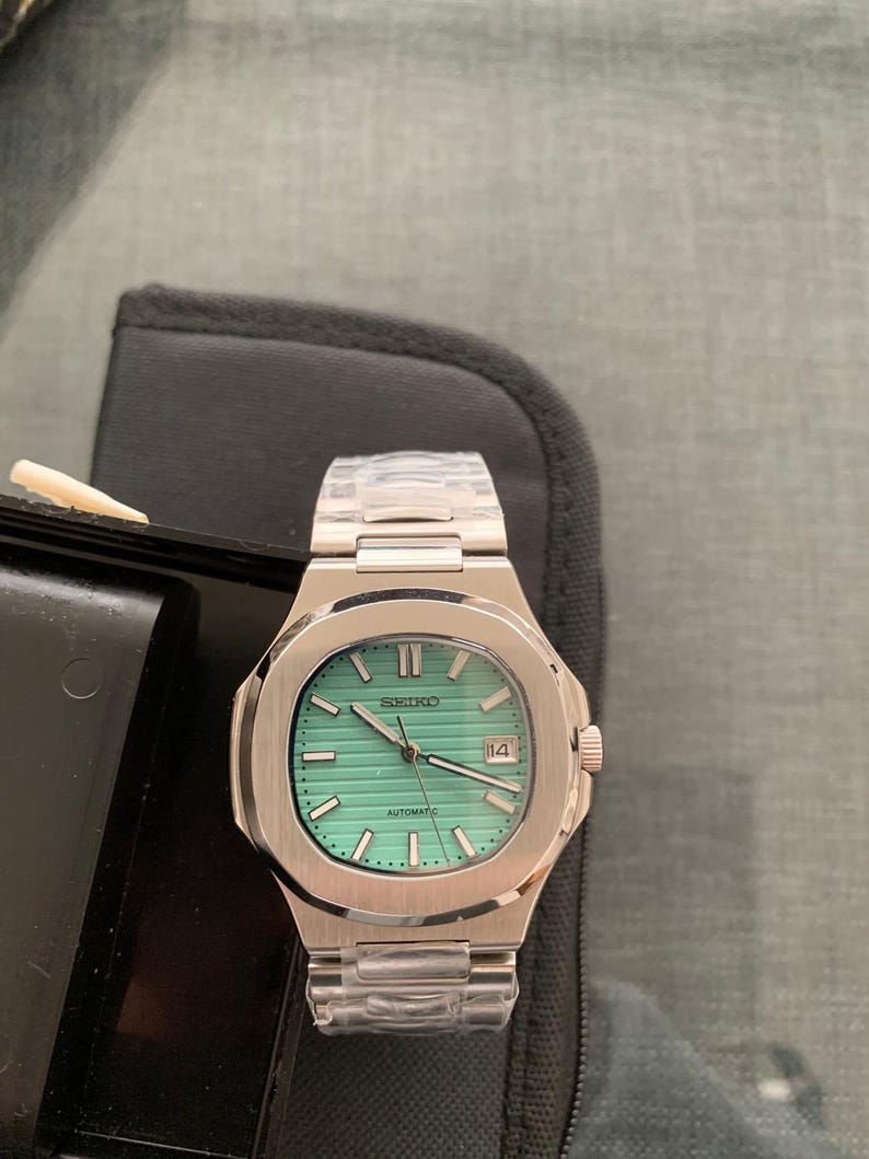May include: A stainless steel Seiko watch with a light green face and a date display at the 3 o'clock position. The watch has a silver band and the word "SEIKO" is printed at the top of the watch face.