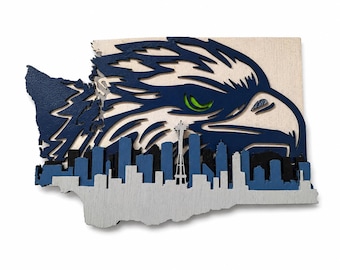 May include: A multi-layered wooden wall art piece shaped like the state of Washington. It features a large eagle head in navy blue and white with a bright green eye, layered over a dark blue and light grey Seattle skyline silhouette including the Space Needle.