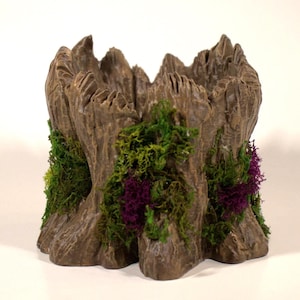 Może przedstawiać: Brązowa donica w kształcie pnia drzewa o szorstkiej, teksturowanej powierzchni. Zielone i fioletowe akcenty mchu zdobią boki donicy. Donica ma wydrążony środek, idealny do przechowywania małych roślin lub przedmiotów dekoracyjnych.