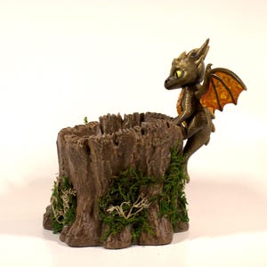 May include: A bronze dragon figure with orange wings perches on a brown tree stump planter. The planter is detailed with green moss, creating a fantasy-themed decorative item. The dragon has yellow eyes.