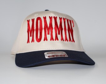 Pode incluir: Boné de beisebol com coroa creme e aba azul marinho. A palavra "MOMMIN'" é bordada em vermelho na frente. Uma pequena etiqueta é visível na aba.