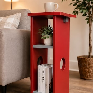 Peut inclure: Une table d'appoint rouge avec deux étagères grises et une surface supérieure. Une tasse blanche est posée sur le dessus, et une petite plante en pot et des livres sont sur les étagères. La table a deux découpes circulaires sur le côté.