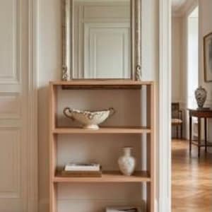 Op de afbeelding: Een licht houten boekenplank met drie open schappen. Het bovenste schap bevat een decoratieve witte kom, het middelste schap bevat een stapel boeken en een witte vaas, en het onderste schap bevat een stapel boeken. Boven de plank hangt een spiegel.