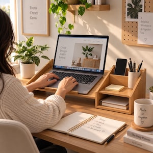 Puede incluir: Un soporte para portátil de madera clara con un organizador de escritorio integrado que cuenta con varios compartimentos para bolígrafos, un smartphone y artículos de papelería, sobre un escritorio con portátil, cuaderno y taza.