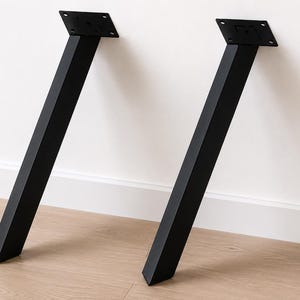 Könnte beinhalten: Zwei mattschwarze Möbelbeine aus Metall mit quadratischem Profil und abgewinkeltem Design. Jedes Bein verfügt über eine flache, rechteckige Montageplatte an der Oberseite mit vier vorgebohrten Löchern zur Befestigung an einer Oberfläche.