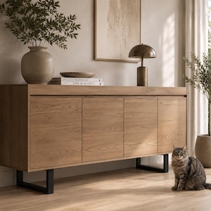 Peut inclure: Un buffet en bois de chêne clair doté de quatre portes d'armoire avec un motif de grain de bois visible, soutenu par deux pieds en métal noir en forme de U. Le buffet est surmonté d'un grand vase en céramique, d'un bol en bois et d'une lampe.