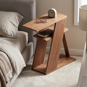 Peut inclure: Une table d'appoint en bois en forme de Z avec une finition en grain de bois naturel. La table présente une surface supérieure plate, une étagère intermédiaire contenant un livre et une base solide. Une petite horloge analogique et des lunettes.