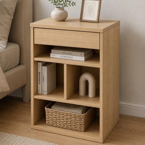 May include: A light oak wood nightstand featuring one top drawer and three open storage compartments. The unit holds a small vase with greenery, a framed print, four books titled Minimalista, KINFOLK, Bauhaus, and Eames, a stone arch, and a woven basket.