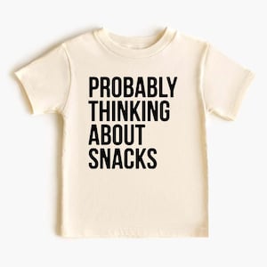 Pode incluir: Camiseta creme com a frase "PROBABLY THINKING ABOUT SNACKS" impressa em letras pretas em negrito. A camiseta de manga curta é feita de um material macio e é adequada para crianças.