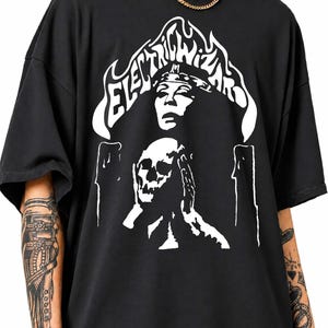 May include: A black short-sleeve t-shirt featuring a white graphic print. The design displays the text Electric Wizard in a stylized font above a figure wearing a headband, positioned over a skull held by two hands, flanked by two candles.
