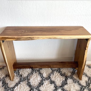 May include: A rustic wooden console table with a live edge top and a lower shelf. The table is made of natural wood with a warm brown color and a natural wood grain pattern. The table is sitting on a white and gray rug.