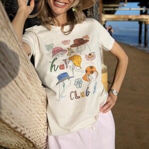 Puede incluir: Camiseta color crema con un gráfico colorido de "Hat Club". El diseño presenta ilustraciones de varios sombreros y elementos florales. La camiseta se combina con pantalones a rayas rosas, creando un look casual y veraniego.