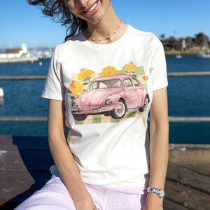 Puede incluir: Camiseta blanca con una ilustración en acuarela de un coche rosa vintage y flores amarillas. El coche está sobre un fondo a cuadros verde y blanco. El diseño incluye pequeños corazones naranjas.