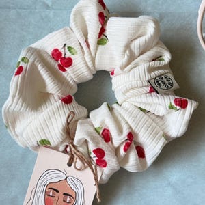Könnte beinhalten: Cremefarbene Scrunchie mit gerippter Textur, verziert mit roten Kirsch- und grünen Blattmustern. Ein Anhänger mit einer Frauenillustration ist mit Bindfaden befestigt. Ein Haaraccessoire.