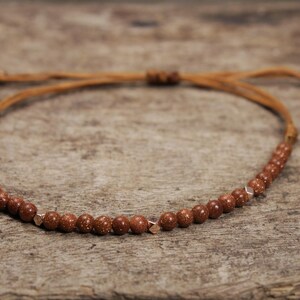 Pode incluir: Um bracelete de cordão de couro marrom com contas de pedra marrom e detalhes dourados.