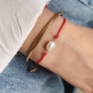 Puede incluir: Un brazalete de cuerda roja con un dije de perla blanca y un brazalete de oro.