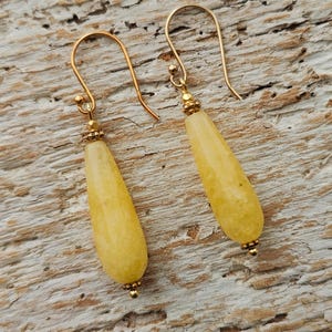 May include: A pair of gold-colored drop earrings with teardrop-shaped, yellow-colored beads. The earrings have a simple design with a hook closure and small gold accents. The earrings are displayed on a light brown, textured wooden surface.