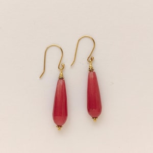 May include: A pair of drop earrings with a gold-colored hook and small gold beads. The earrings feature a faceted, teardrop-shaped, red-pink stone. The earrings are displayed against a white background.