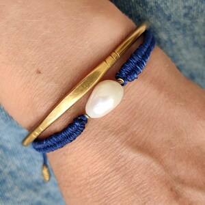 Puede incluir: Un brazalete de oro con un brazalete de cordón trenzado azul superpuesto. Una perla blanca está unida al cordón trenzado.