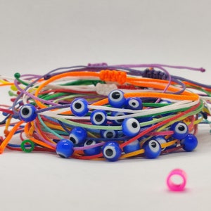 Puede incluir: Un montón de pulseras de cuerda de colores con cuentas azules de ojo turco. Las pulseras están hechas de varios colores, incluyendo naranja, blanco, morado y verde.