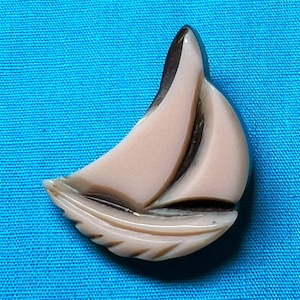May include: A vintage sailboat brooch in shades of tan and brown, set against a blue background. The brooch features a detailed design of a sailboat with a textured sail and hull. The brooch is likely made of plastic or resin.