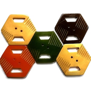 May include: A collection of five hexagonal buttons in yellow, dark brown, dark green, and orange. Each button features two central holes, two rectangular cutouts, and textured parallel lines along two of the six sides of the hexagon shape.