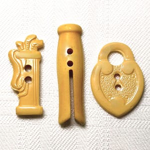 May include: Three vintage yellow buttons: a golf bag, a clothespin, and a heart-shaped padlock. Each button has holes for sewing. The golf bag button features golf clubs. The clothespin button has a split design. The padlock button has a textured surface.