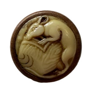 May include: A round, off-white button with a detailed carving of a rat curled around a stylized object. The rat is cream-colored, and the button has a dark brown, textured border. The design is raised, creating a three-dimensional effect.