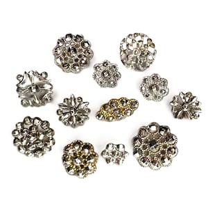 May include: A collection of eleven decorative buttons in various shapes and sizes, featuring metallic silver and gold finishes with faceted, reflective surfaces. The buttons include circular, floral, and oval designs with intricate textures and patterns.