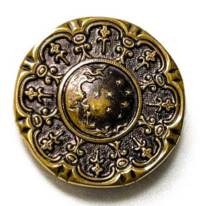 May include: A round, antique brass button with an ornate design. The center features a crescent moon and stars. The outer edge has a decorative border with floral and cross-like elements. The button has a vintage, celestial theme.