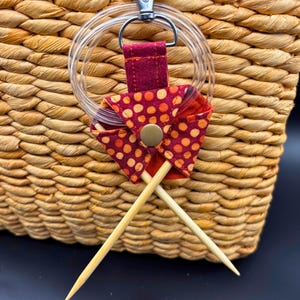 May include: A knitting needle holder with a clear plastic ring and a burgundy fabric accent featuring orange polka dots. Two wooden knitting needles are held in place by the fabric. The holder is attached to a woven basket.