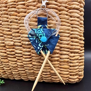 May include: A woven basket with a blue and green patterned knitting needle holder. The holder is attached to a silver clip and holds two wooden knitting needles. The holder has a blue button closure and a clear circular ring.