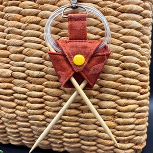 May include: Close-up of a woven basket with a red knitting needle holder. The holder has a yellow button and a clear ring attached. Two wooden knitting needles are inserted into the holder. The basket is made of natural fibers.