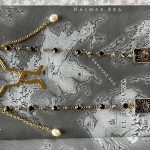 May include: Gold bookmark with a distinctive shape, accompanied by two chains embellished with black beads and pearls. It's placed on a map-style background with text, adding a touch of elegance.