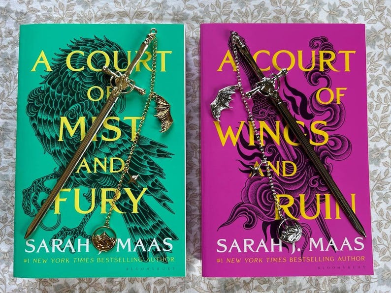 May include: Two books with decorative sword-shaped bookmarks. The book on the left has a teal cover with the title "A Court of Mist and Fury" and a bird illustration. The book on the right has a pink cover with the title "A Court of Wings and Ruin".