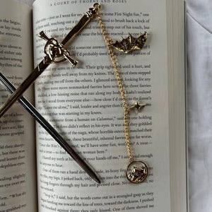 May include: Gold-coloured metal sword and bat-shaped bookmarks with chain and star accents, on an open book. The book's title is "A Court of Thorns and Roses."