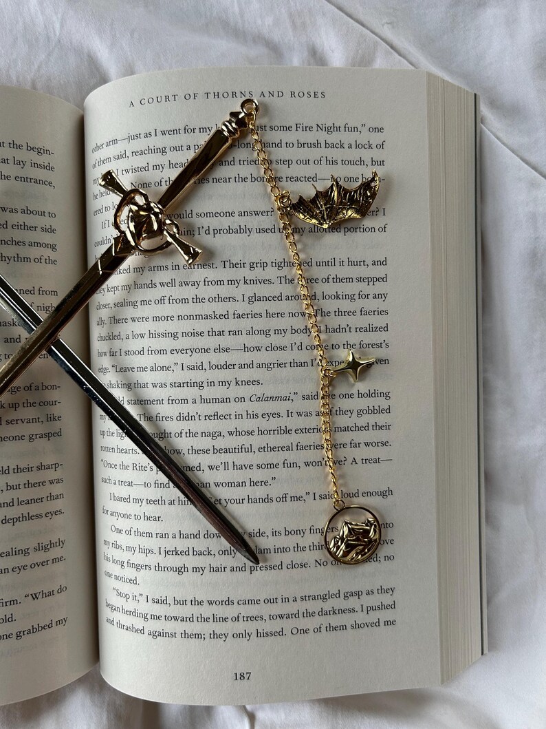 May include: Gold-coloured metal sword and bat-shaped bookmarks with chain and star accents, on an open book. The book's title is "A Court of Thorns and Roses."