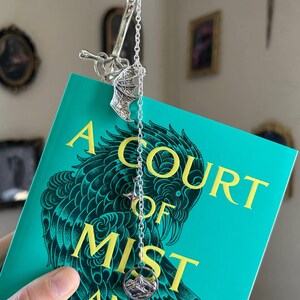 May include: A silver bookmark with a sword and chain design, placed on a book with a teal cover. The book's title, "A Court of Mist and Fury," is in yellow lettering. The bookmark has a mountain charm.
