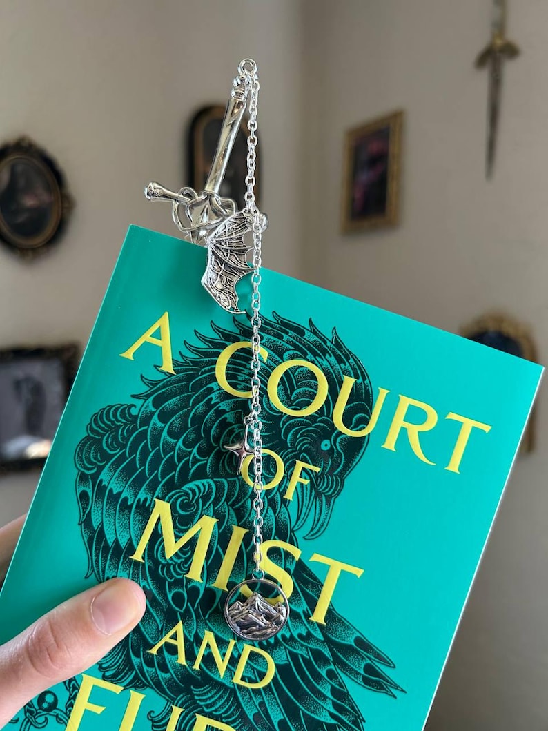 May include: A silver bookmark with a sword and chain design, placed on a book with a teal cover. The book's title, "A Court of Mist and Fury," is in yellow lettering. The bookmark has a mountain charm.