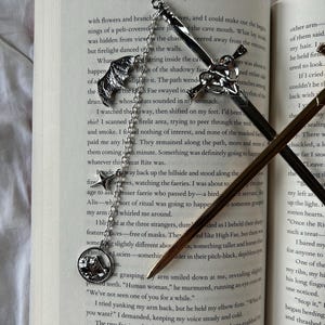 May include: Intricate silver metal bookmarks with decorative charms, including a sword, a wing, and a mountain scene, lie on the open pages of a book. The bookmarks are detailed, adding a touch of fantasy to the reading experience.