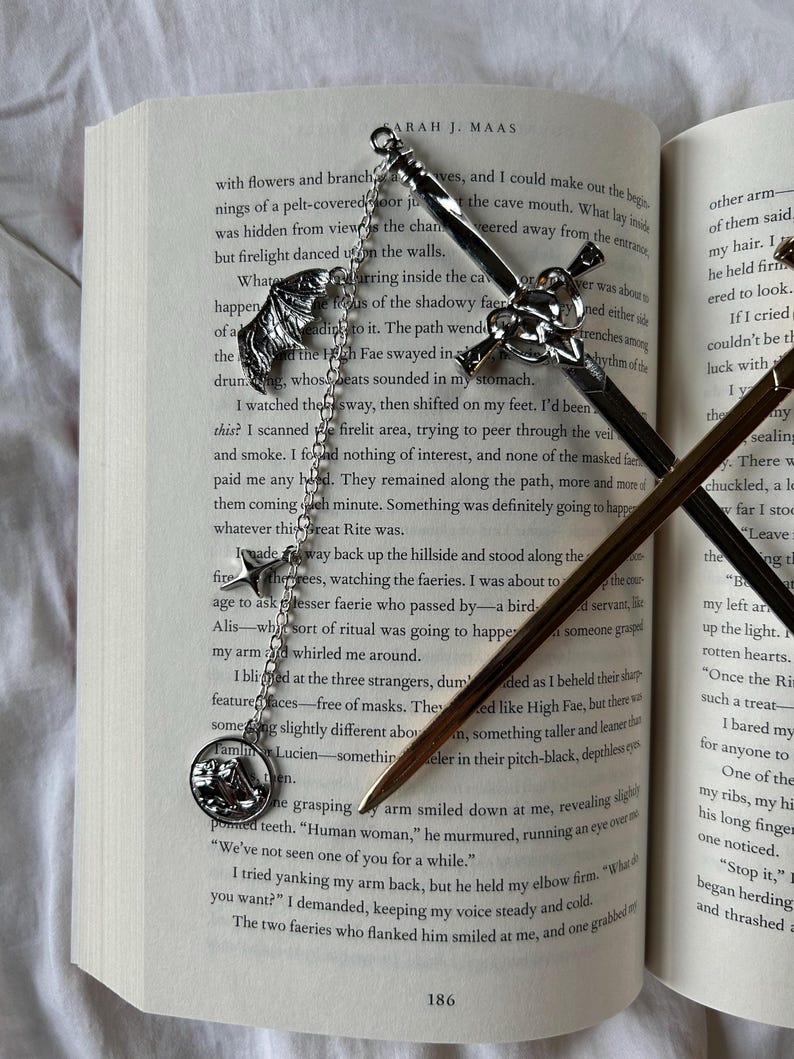 May include: Intricate silver metal bookmarks with decorative charms, including a sword, a wing, and a mountain scene, lie on the open pages of a book. The bookmarks are detailed, adding a touch of fantasy to the reading experience.
