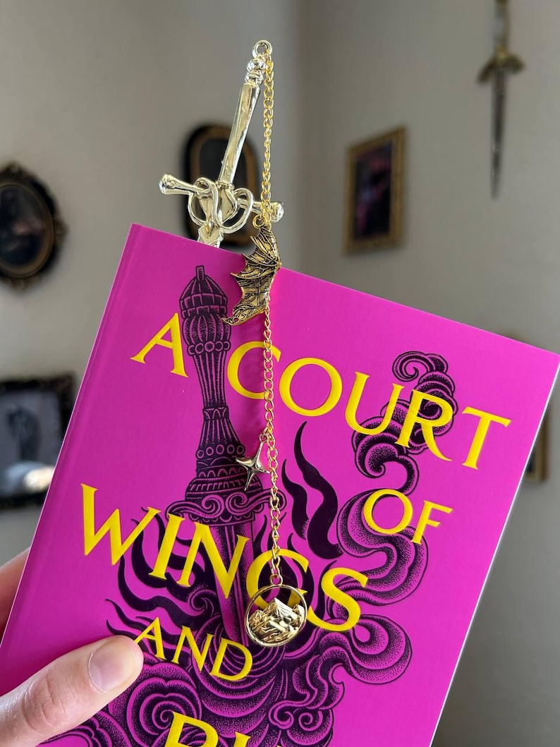 May include: A gold-coloured metal bookmark with a sword design, bat charm, star charm, and a circular charm with a mountain scene. The bookmark is attached to a book with a pink cover and the title "A Court of Wings and Ruin".