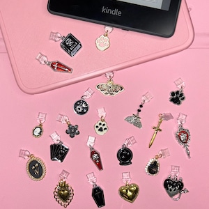 Op de afbeelding: Een verzameling decoratieve bedels met verschillende ontwerpen, waaronder een kist, pootafdrukken, een mot en een hart. De bedels zijn bevestigd aan doorzichtige clips en worden gepresenteerd op een roze oppervlak naast een Kindle.